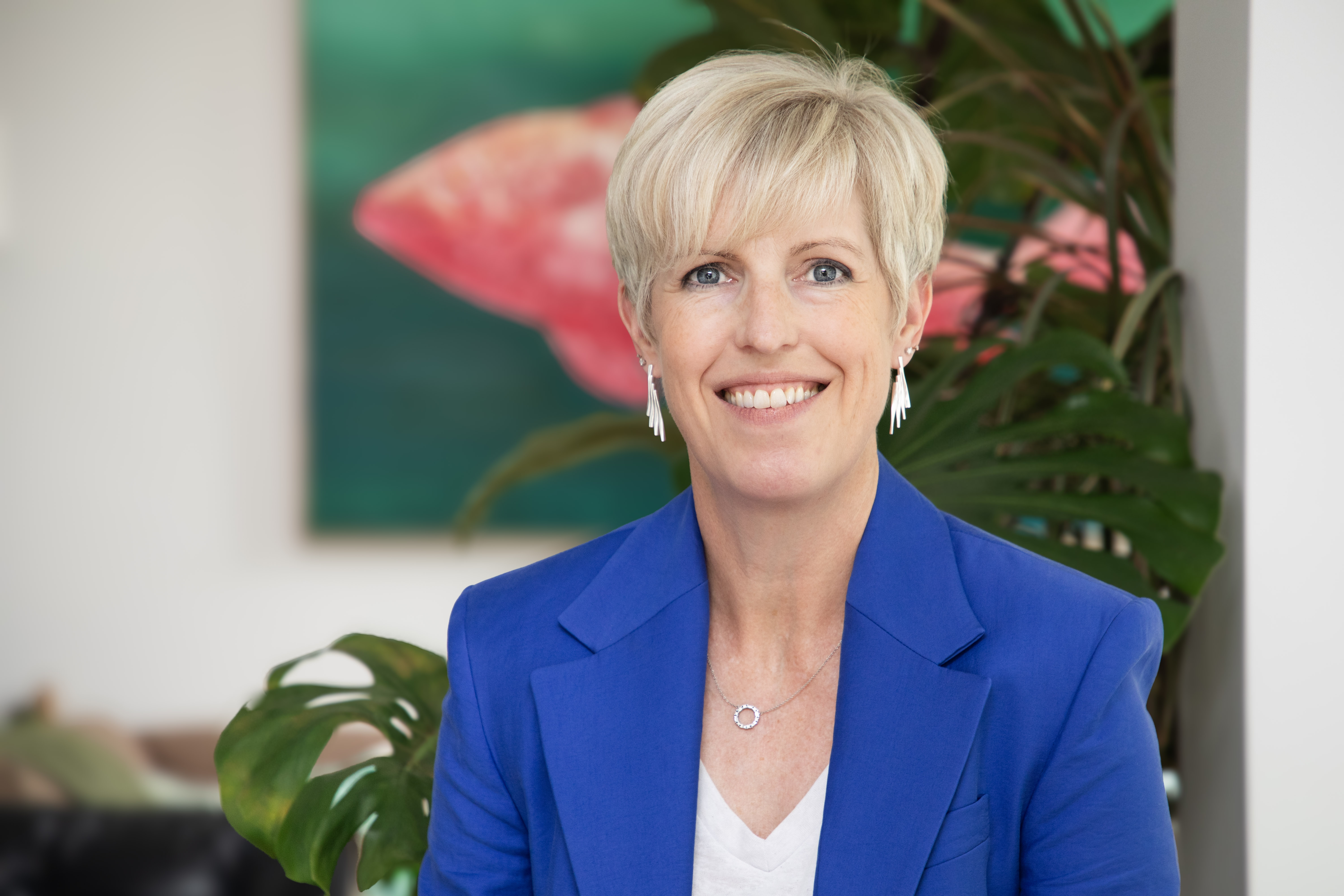 Portrait of Kate, a professional woman with short blonde hair, wearing a blue blazer, smiling against a background of green plants and abstract art.